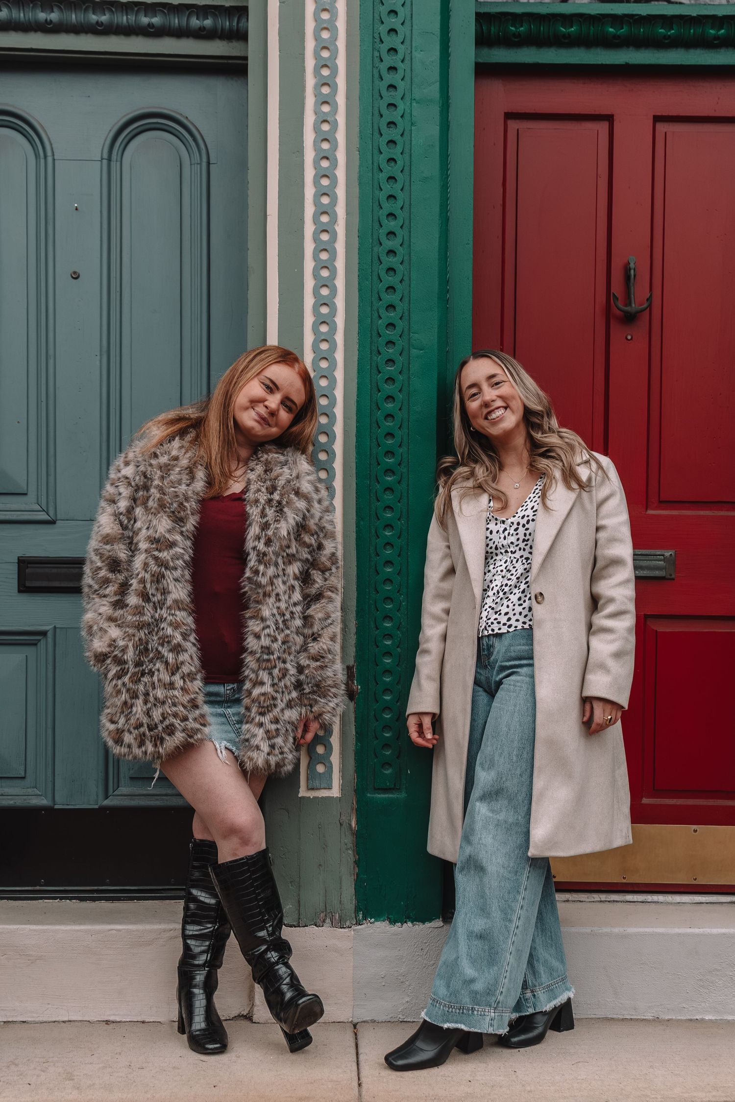 Two women in stylish coats pose smiling against colorful green and red doors on a city street.