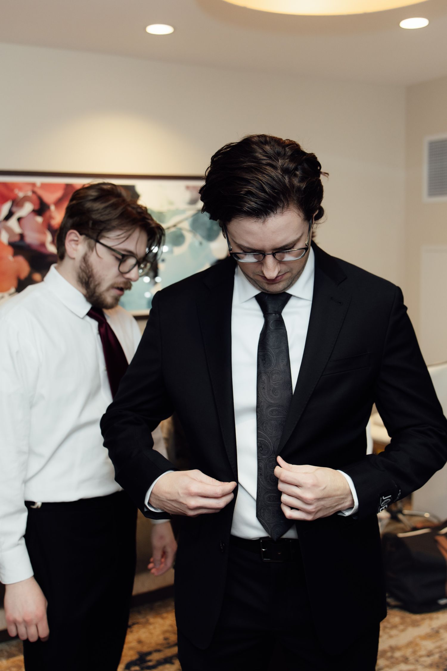 Groom in black suit adjusting tie with groomsman in white shirt and burgundy tie assisting in hotel room.