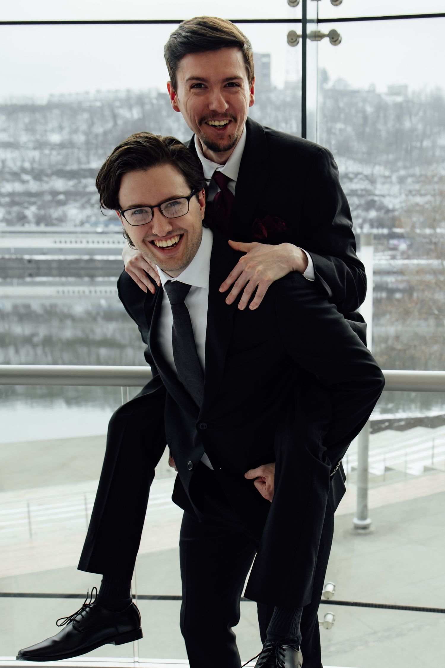 Two men in black suits laughing and doing a piggyback ride indoors with a snowy cityscape visible through glass windows.