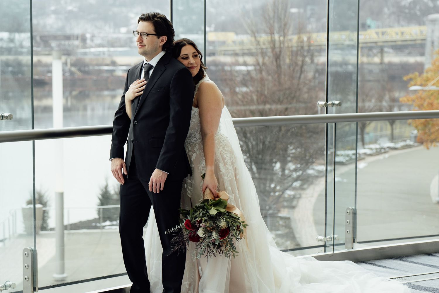 Bride in layered white gown leans against groom in black suit by glass windows overlooking a snowy winter landscape.