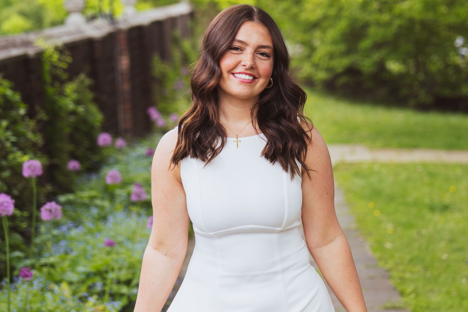 A person in a sleeveless white dress poses outdoors in a garden with blooming flowers and greenery.