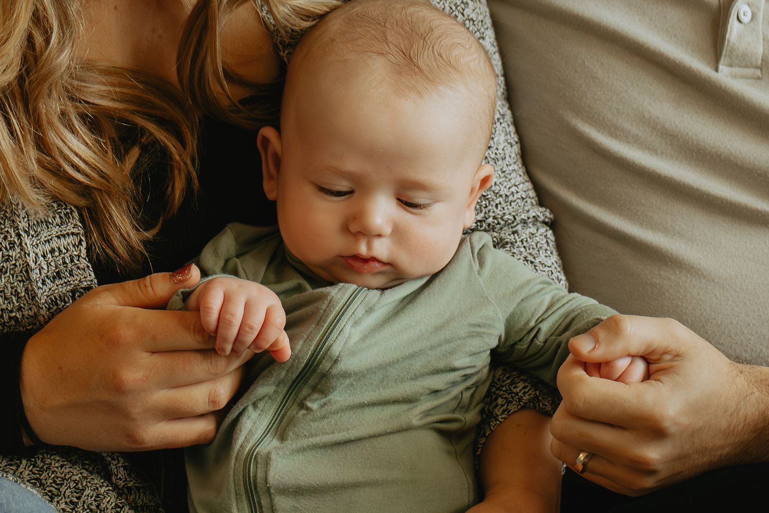 Newborn baby in green onesie resting peacefully between parents, gently held by both mother and father.