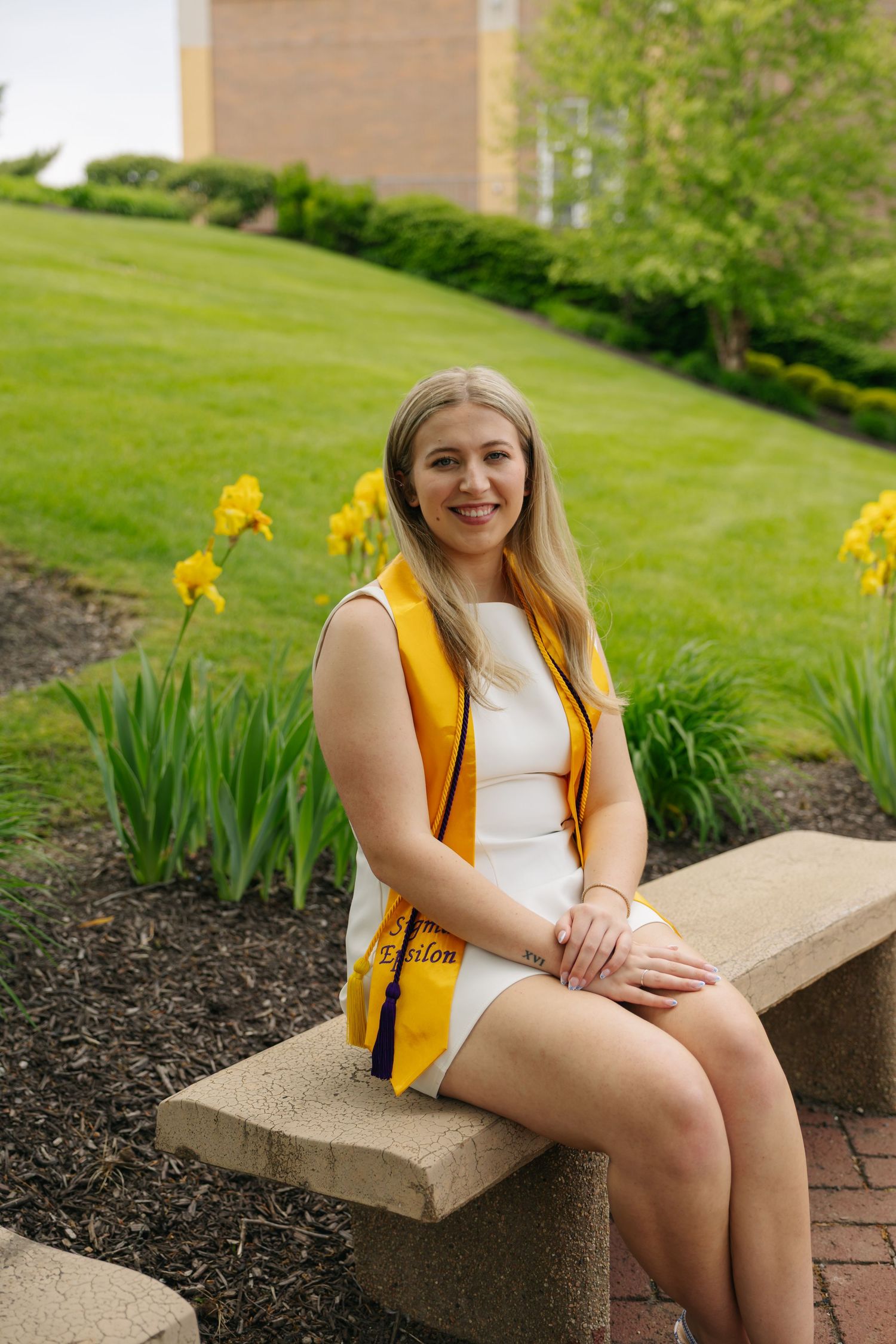 Smiling female graduate in white dress and gold stole sits on stone bench outdoors with yellow flowers.