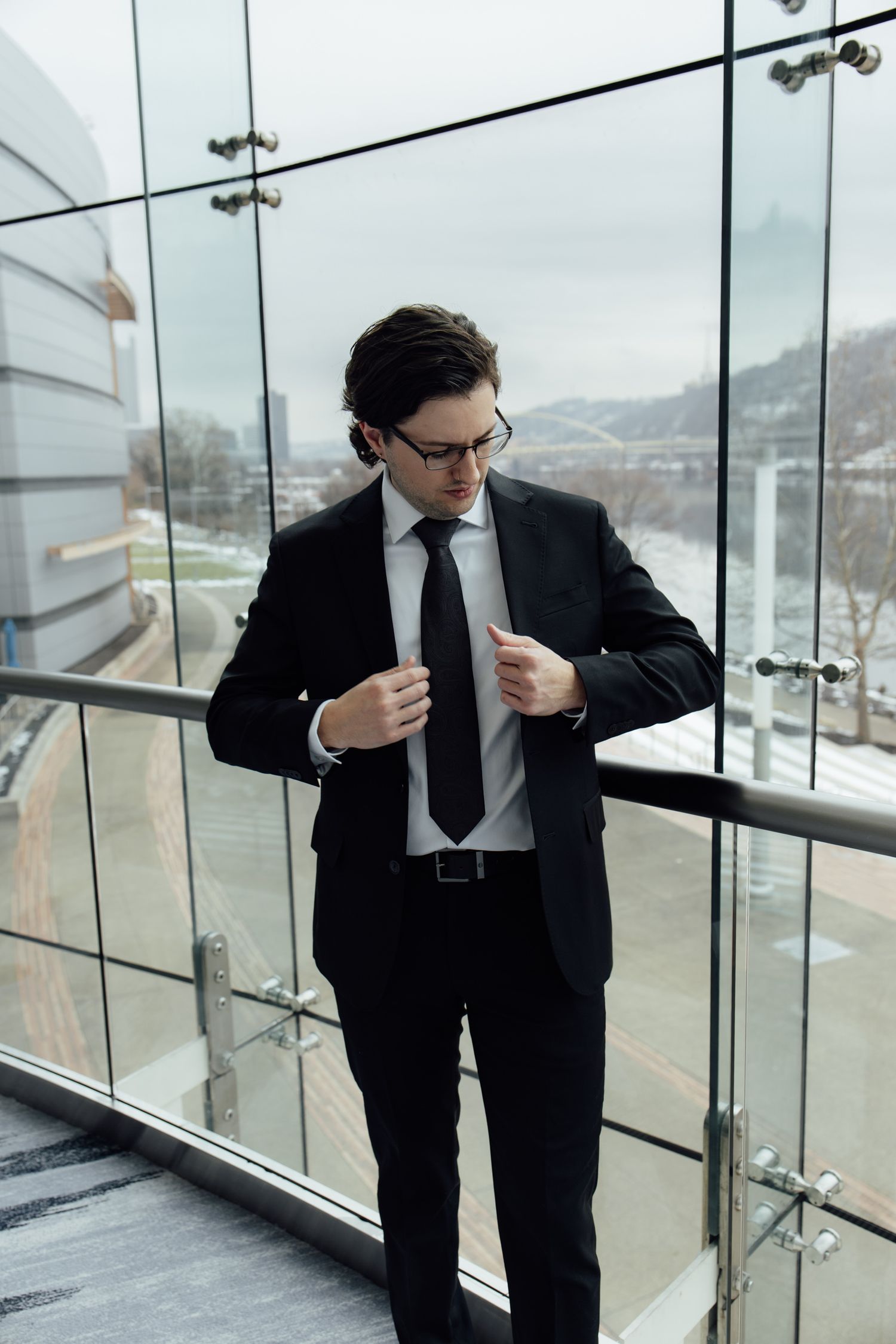 A professional businessman in a black suit and tie adjusting his jacket on a modern glass balcony.
