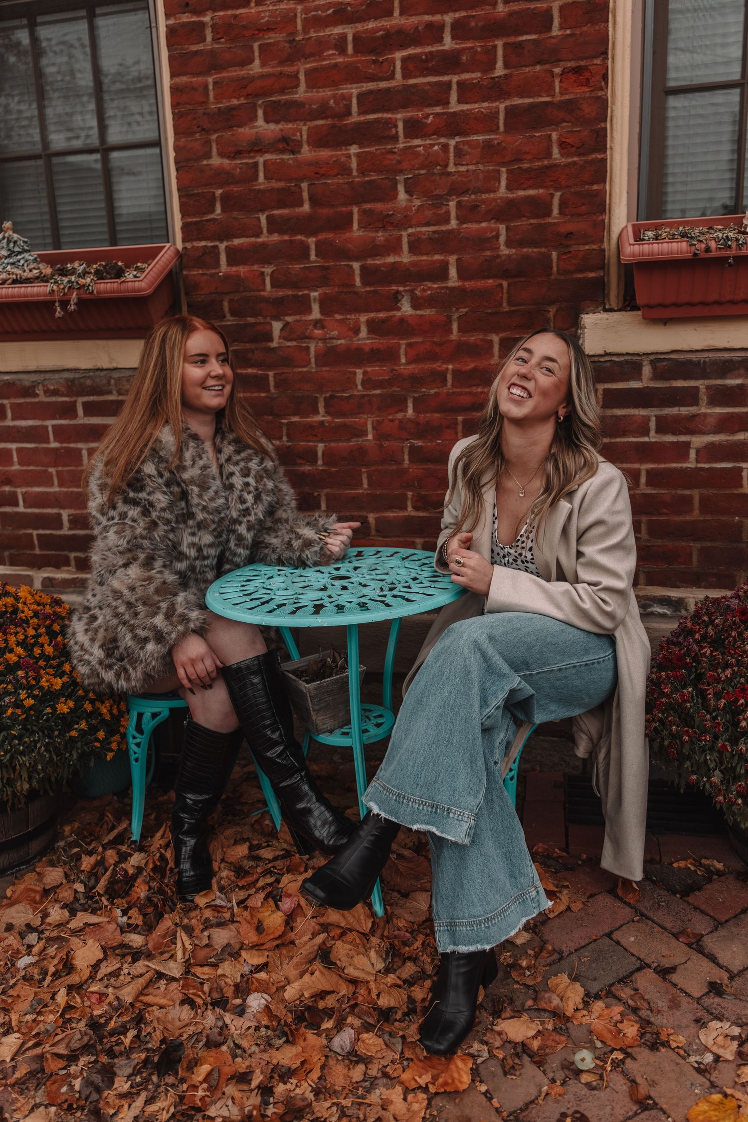 Two women laughing outdoors at a teal table surrounded by fall leaves against a brick wall.