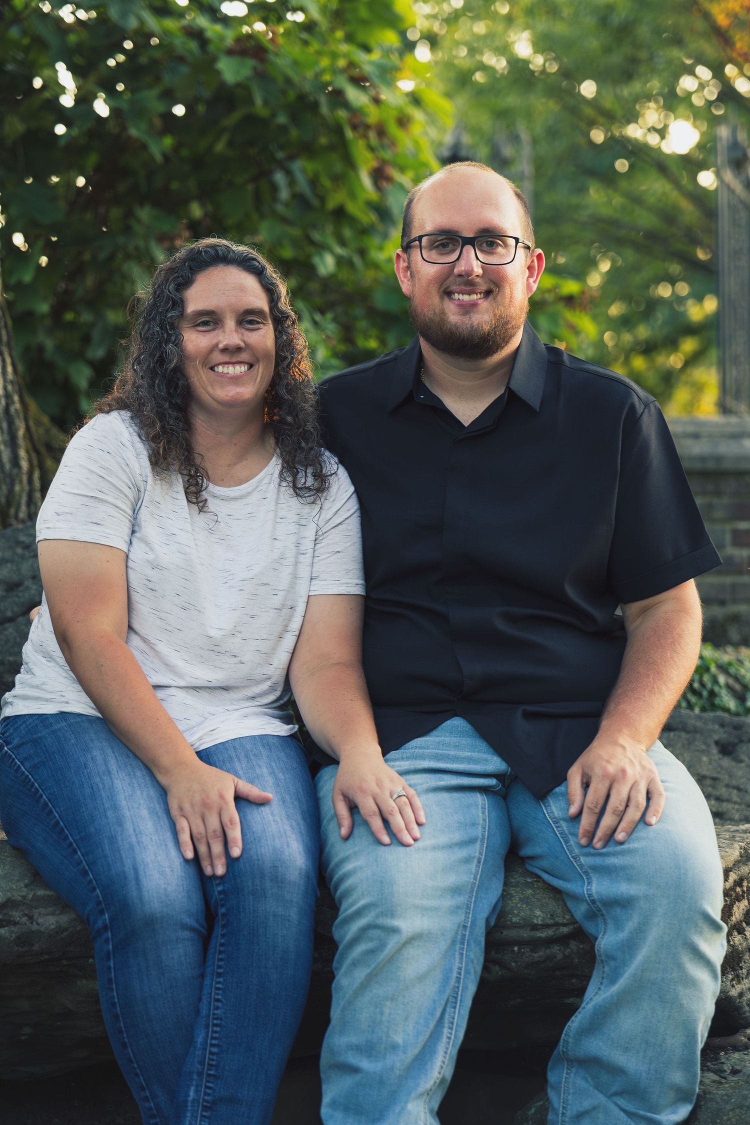 A smiling couple sitting outdoors on a stone ledge surrounded by lush green trees in a warm natural setting.