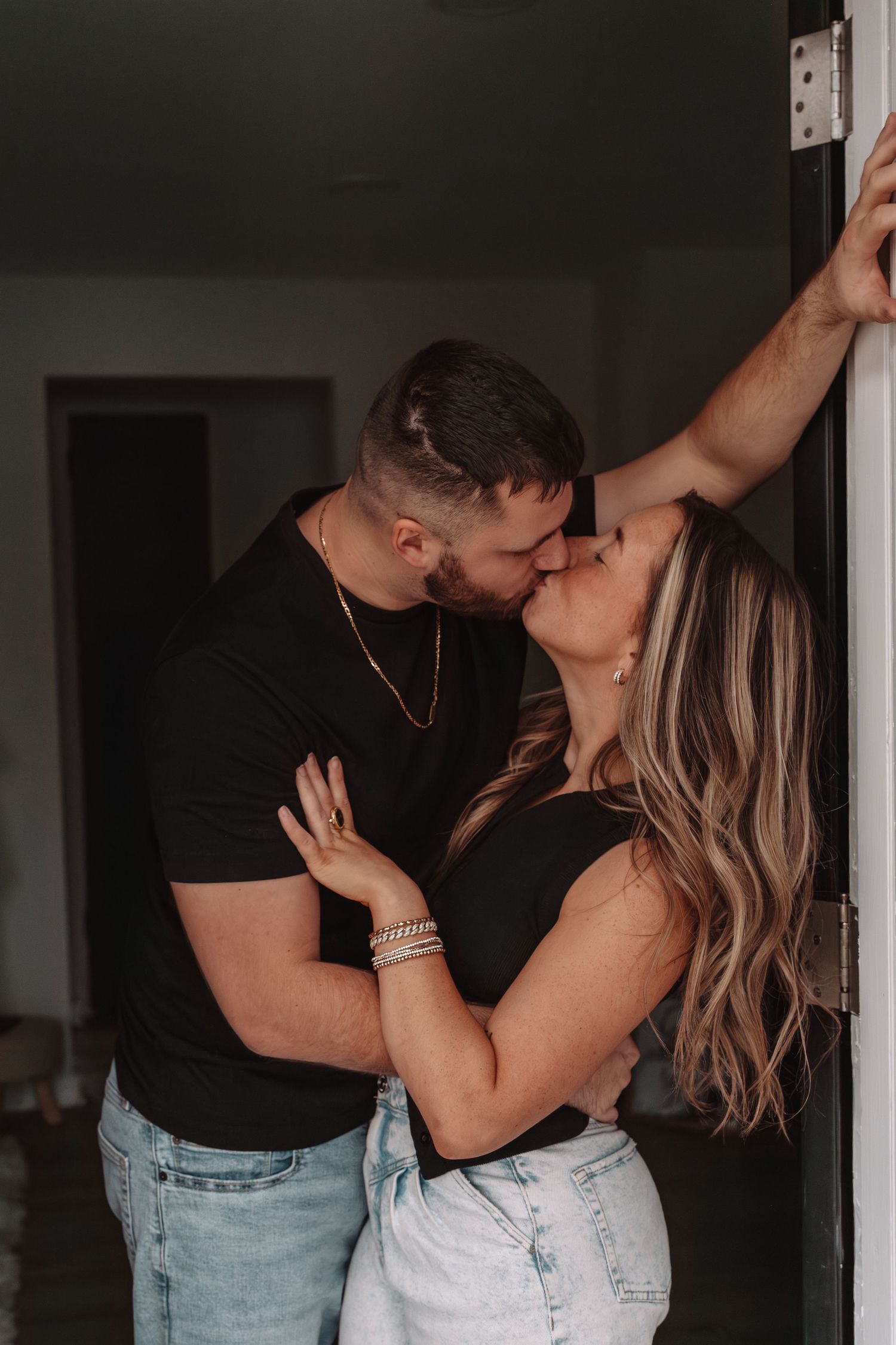 Romantic couple sharing a passionate kiss in a doorway, both wearing black tops and light jeans indoors.