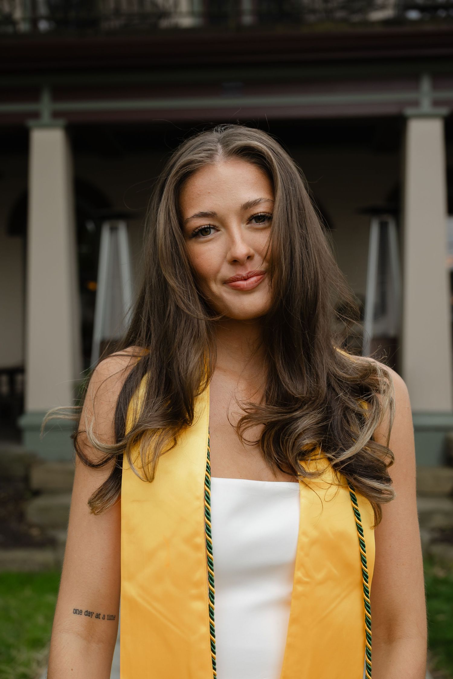 Young woman in white dress and gold graduation stole smiles outdoors at her graduation celebration.