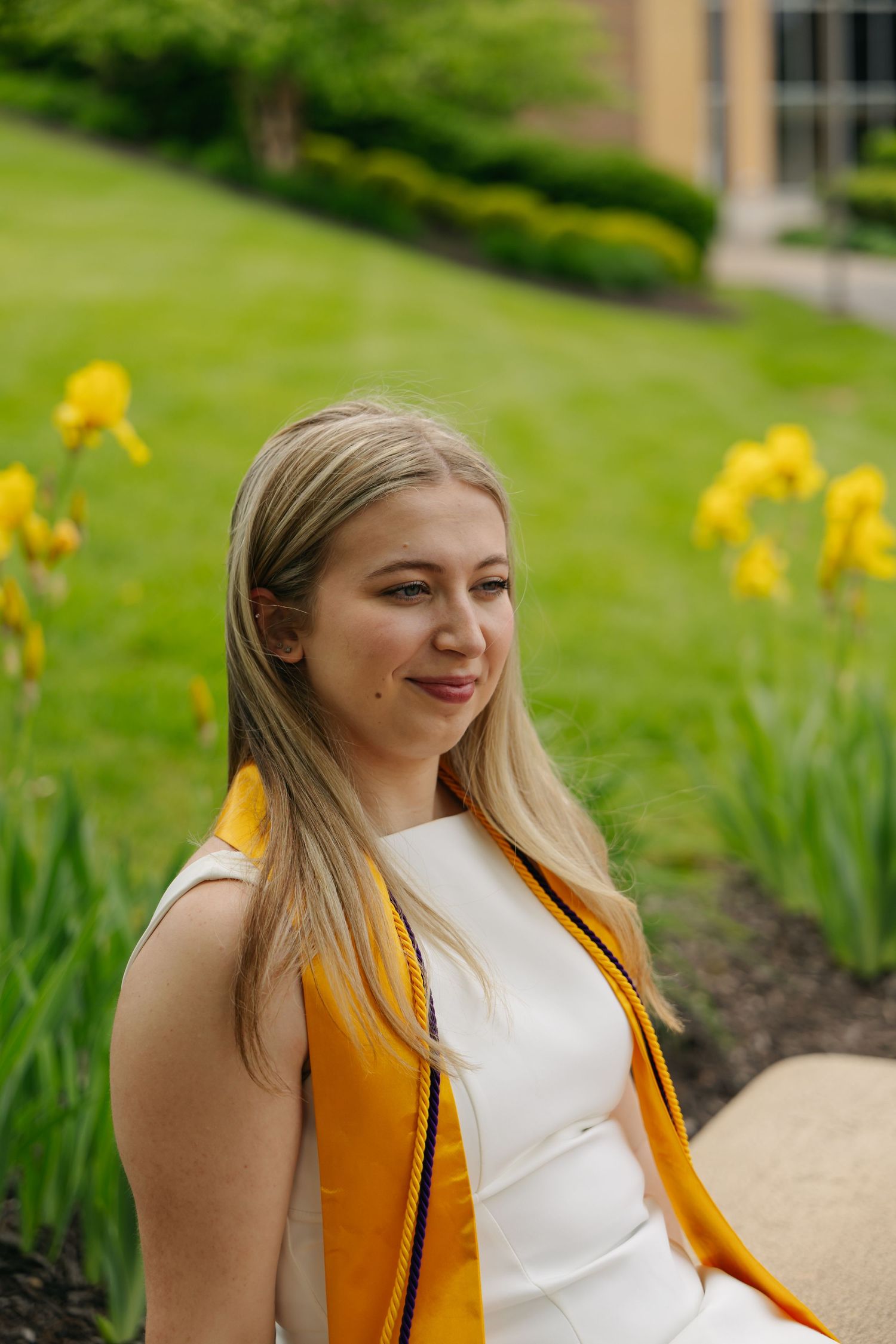 Blonde woman in white dress with gold graduation stole sitting outdoors near yellow iris flowers.