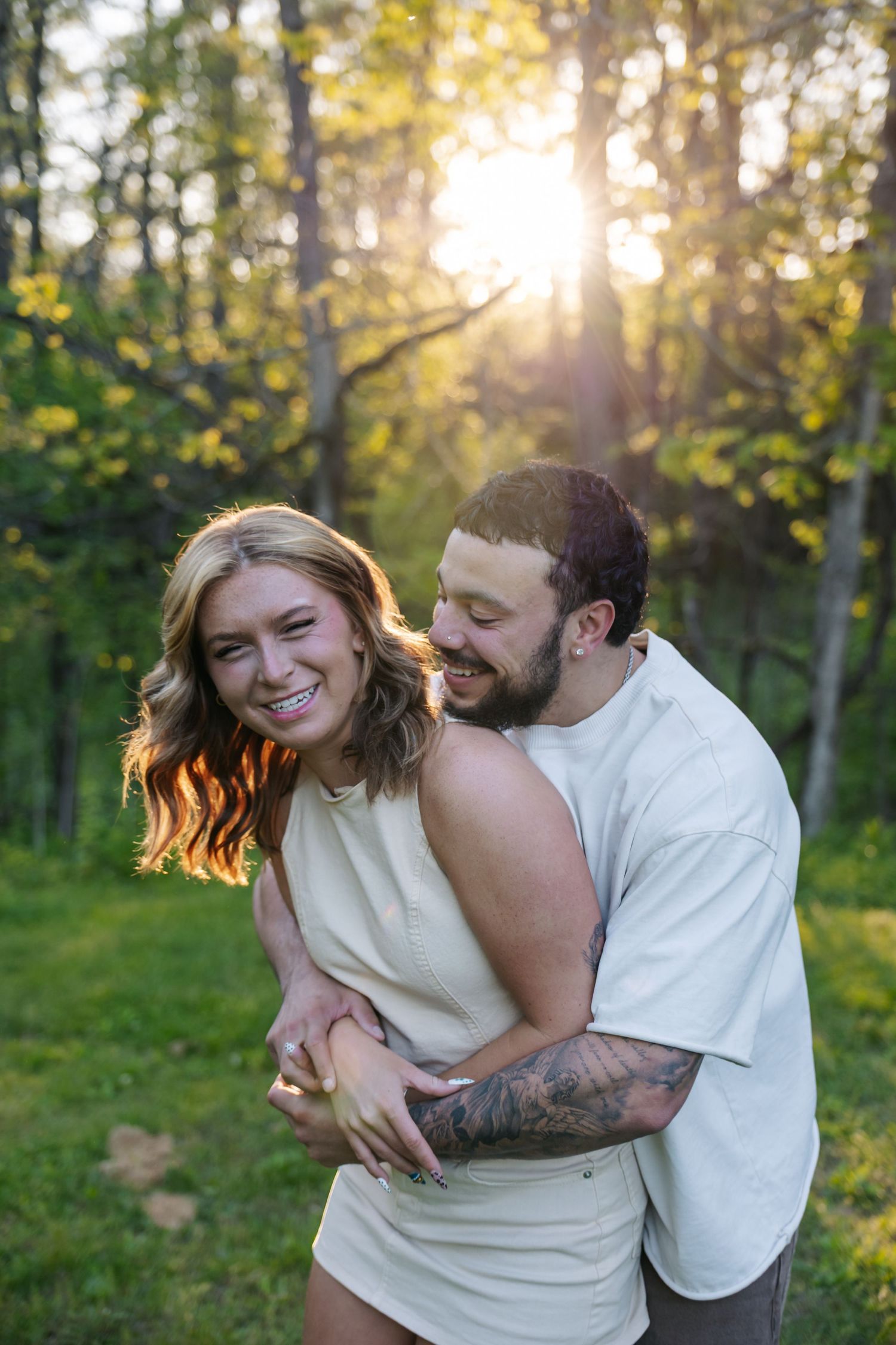 A smiling couple embraces in a sunlit forest, woman in white dress, man in white tee with tattoos, golden hour glow.
