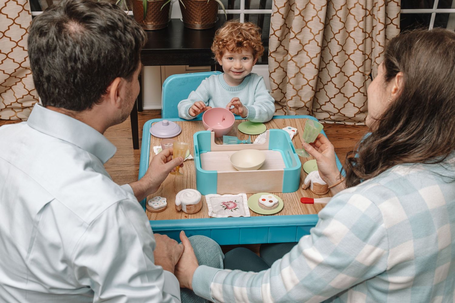 Family playing with toy kitchen set, parents watching toddler serve pretend food at small blue table.
