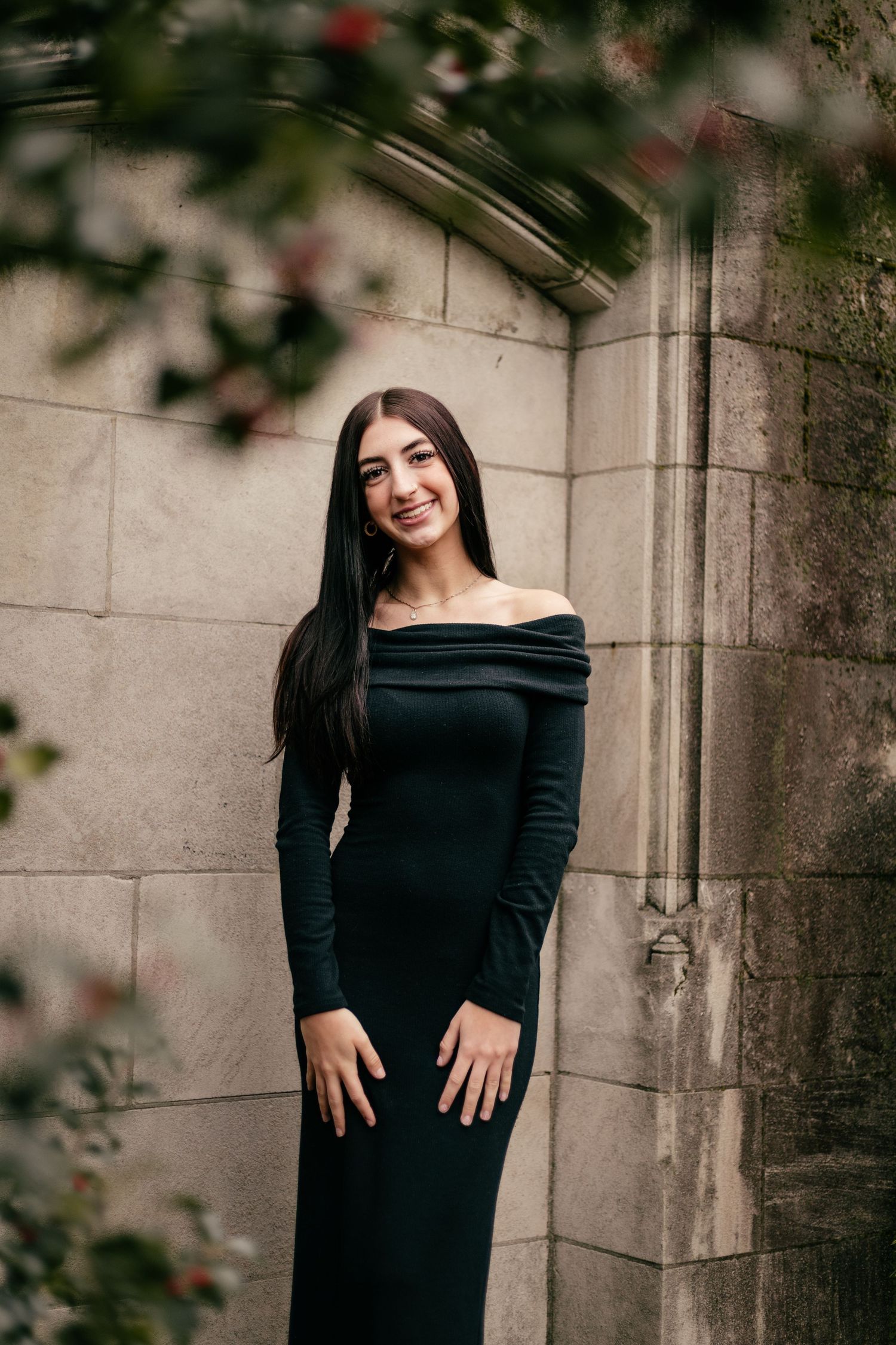 Woman in black off-shoulder gown poses against stone wall with blurred foliage.