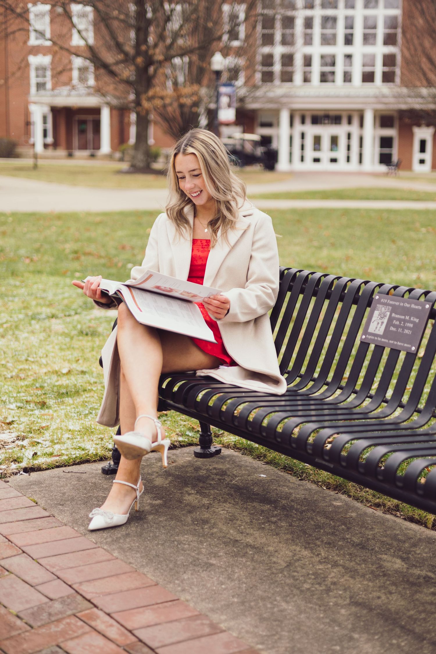 Graduate reading on campus bench wearing cream coat and red dress against brick building.