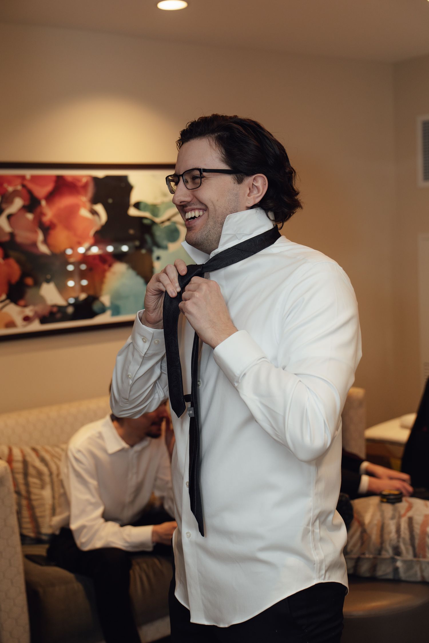 Smiling groom in white shirt adjusting black tie while getting ready for wedding ceremony indoors.