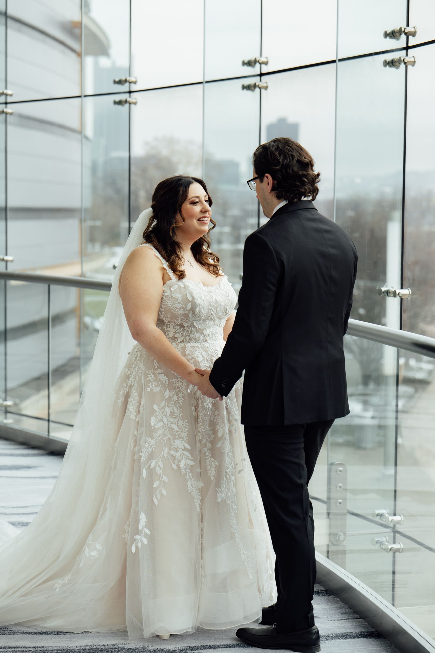 Bride in lace ballgown holds hands with groom in black suit on glass-walled venue balcony.