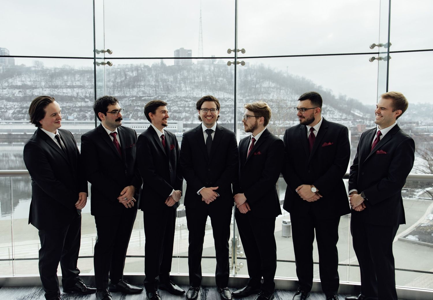 Seven groomsmen in matching black suits laugh together at a modern glass-walled wedding venue in winter.