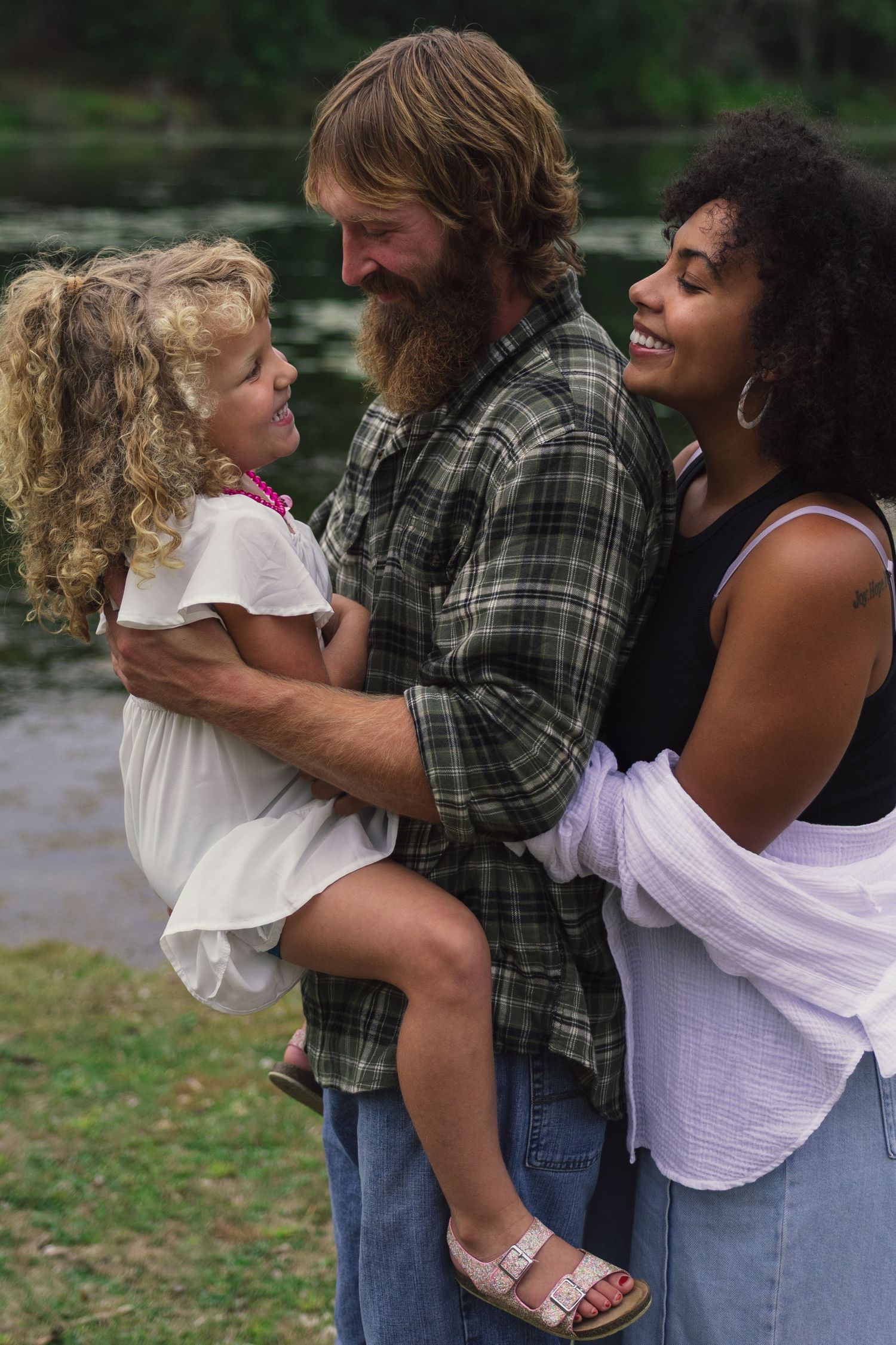 A happy interracial family with a bearded father, curly-haired mother, and young blonde daughter sharing a joyful moment outdoors.