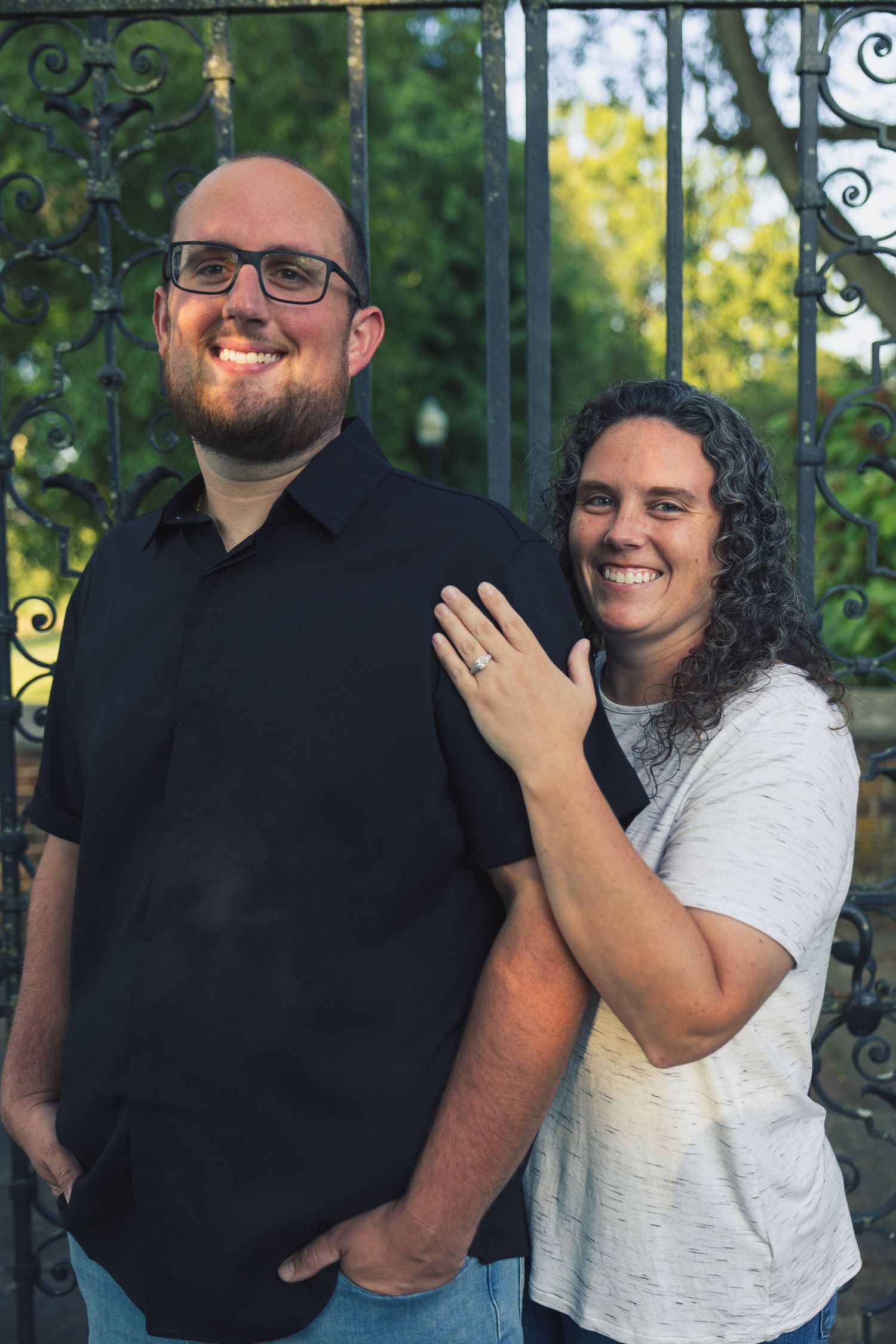 Smiling couple posing outdoors near ornate iron gate, woman with curly hair resting hand on man's shoulder.