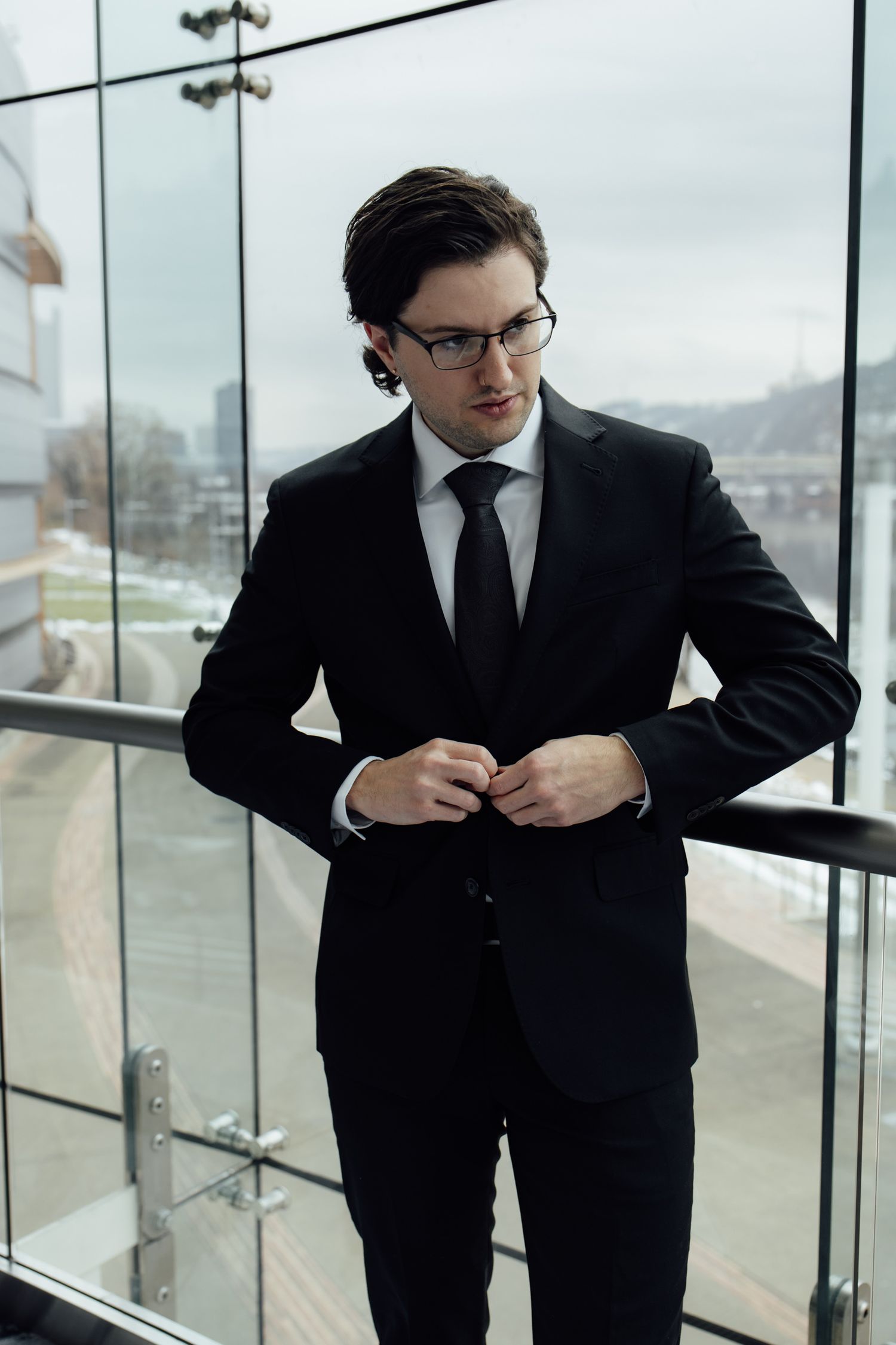 Stylish man in black suit and glasses buttoning jacket on modern glass balcony with city view.