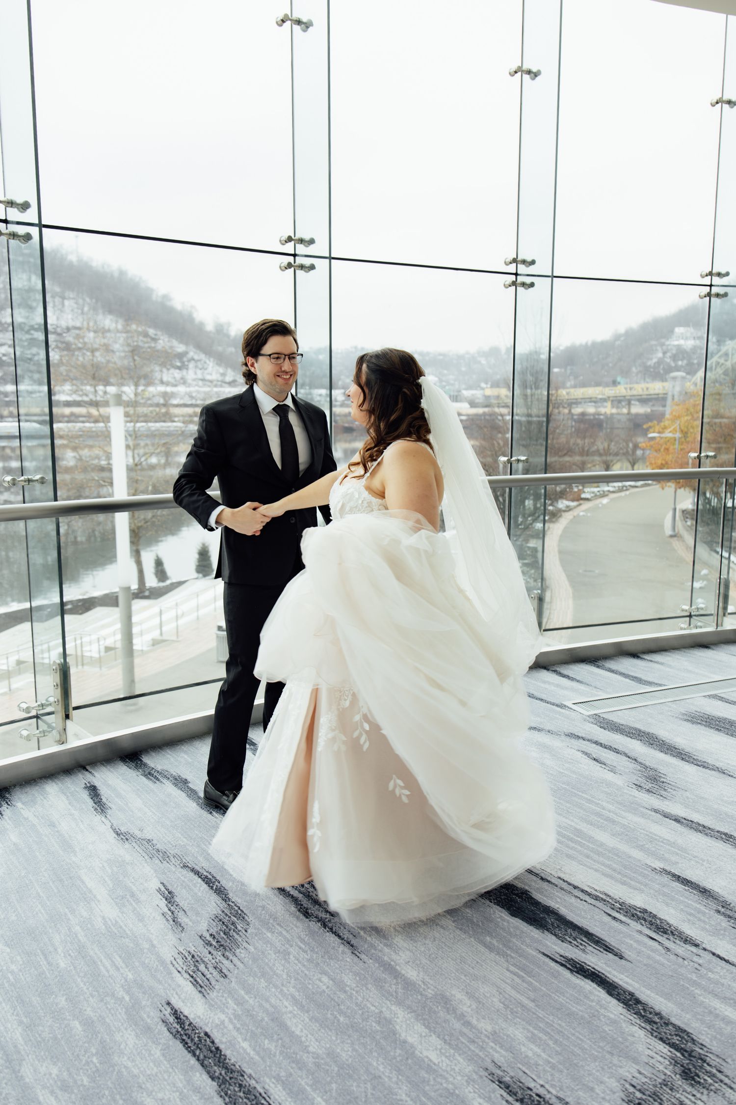 A bride in a flowing white gown and groom in a black suit share a joyful dance near large windows with a winter view.