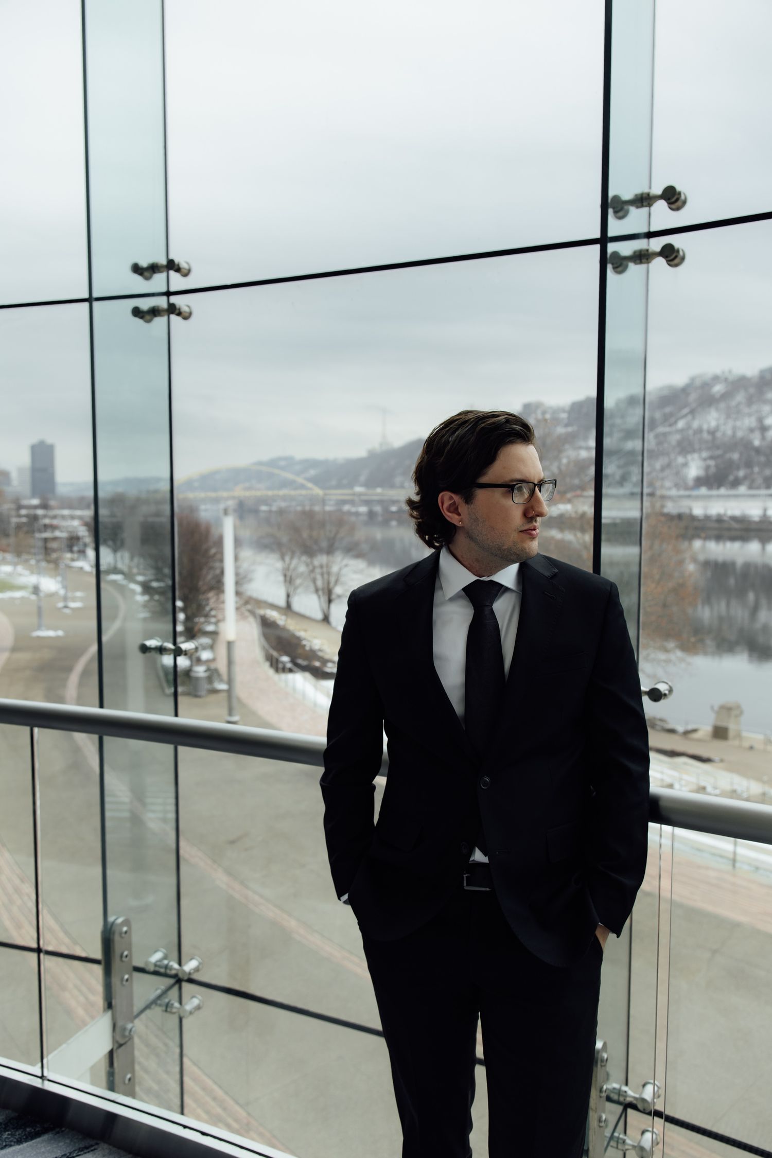 A professional businessman in a dark suit and glasses stands by glass windows overlooking a snowy riverside cityscape.