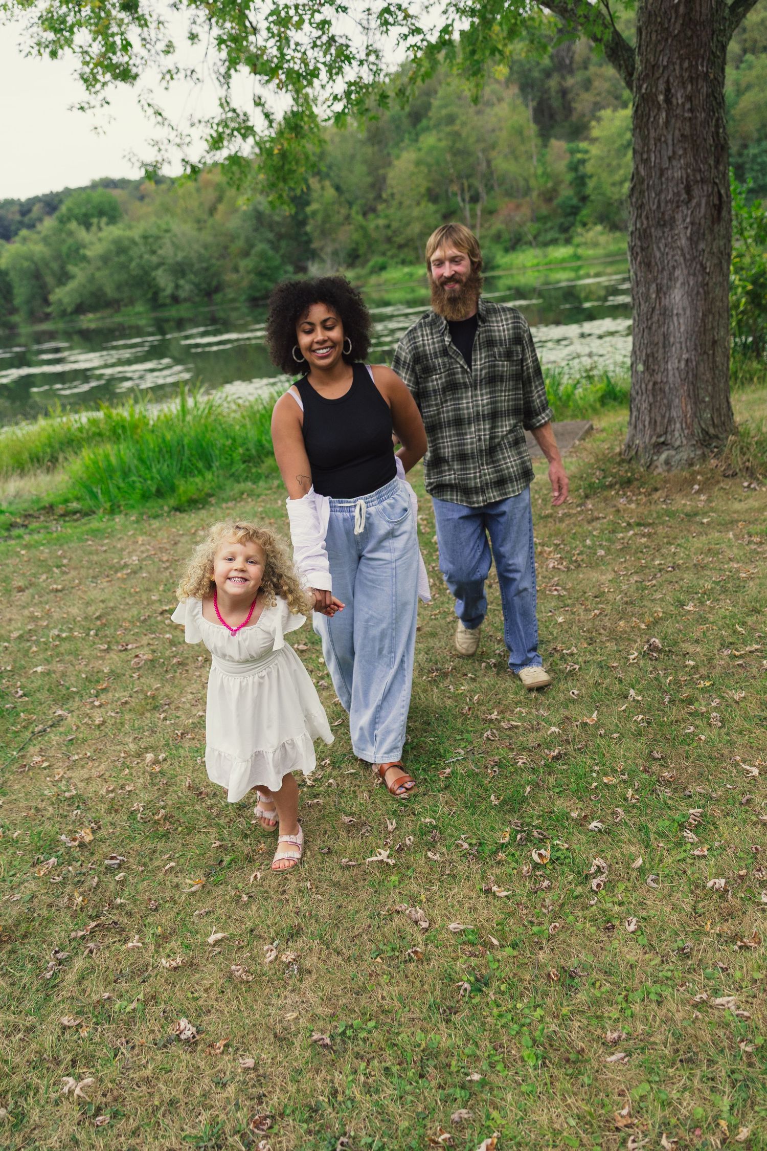 Happy interracial family with curly-haired girl walking by a scenic lake surrounded by lush green trees.