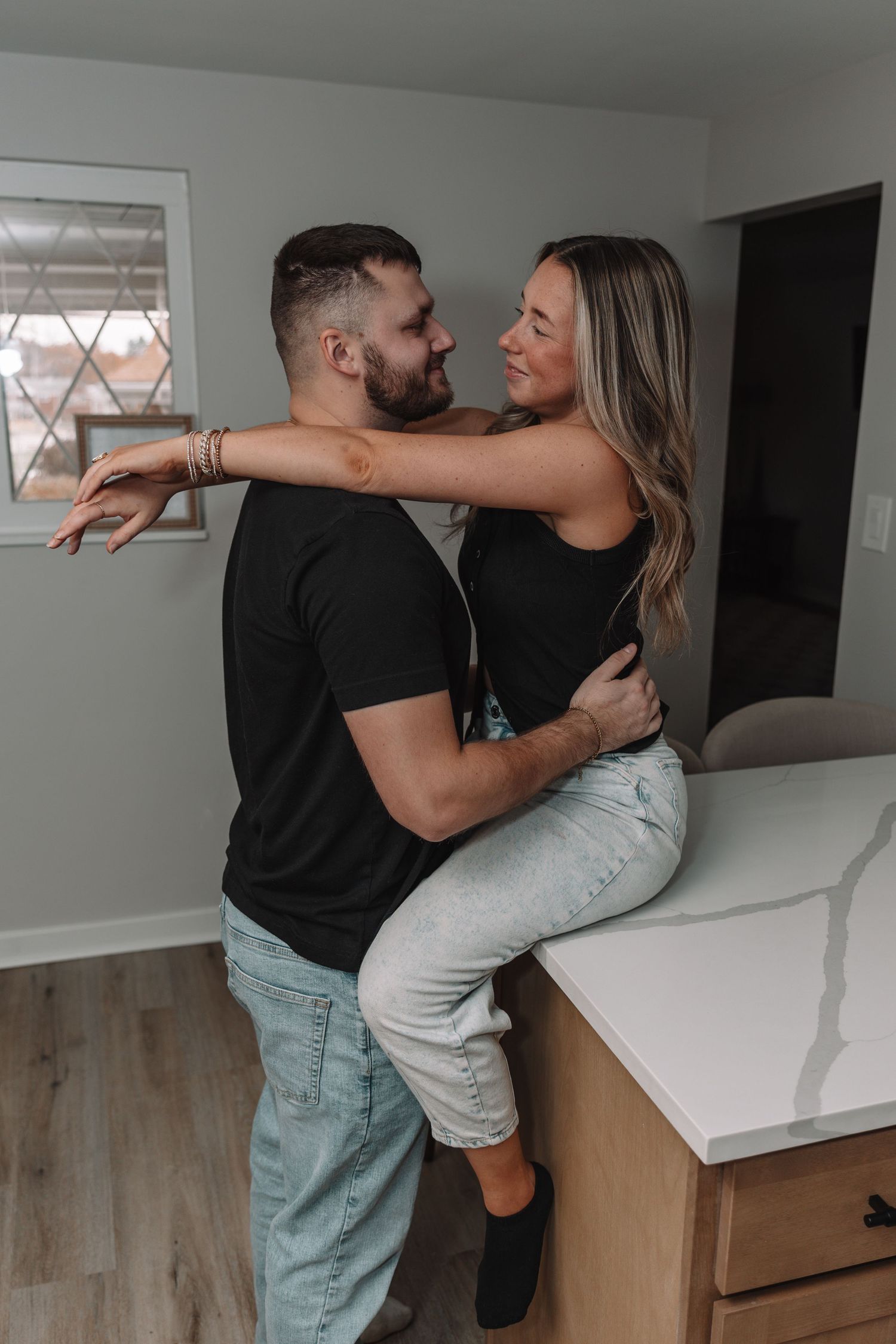 A couple embracing in a kitchen, woman sitting on counter, both wearing black tops and light jeans, smiling at each other.
