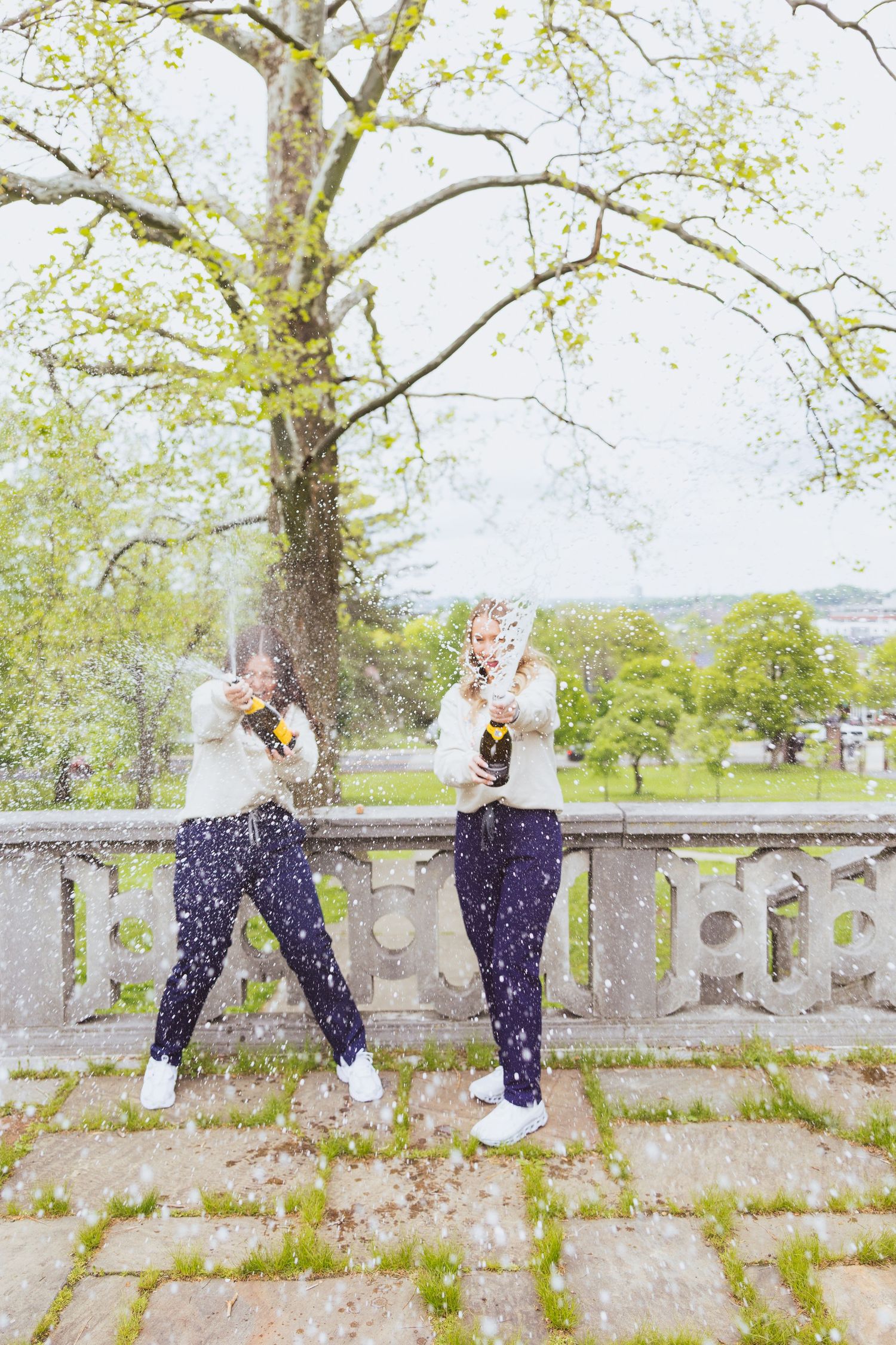 Two graduates celebrate by throwing confetti on stone balcony with green trees in background.