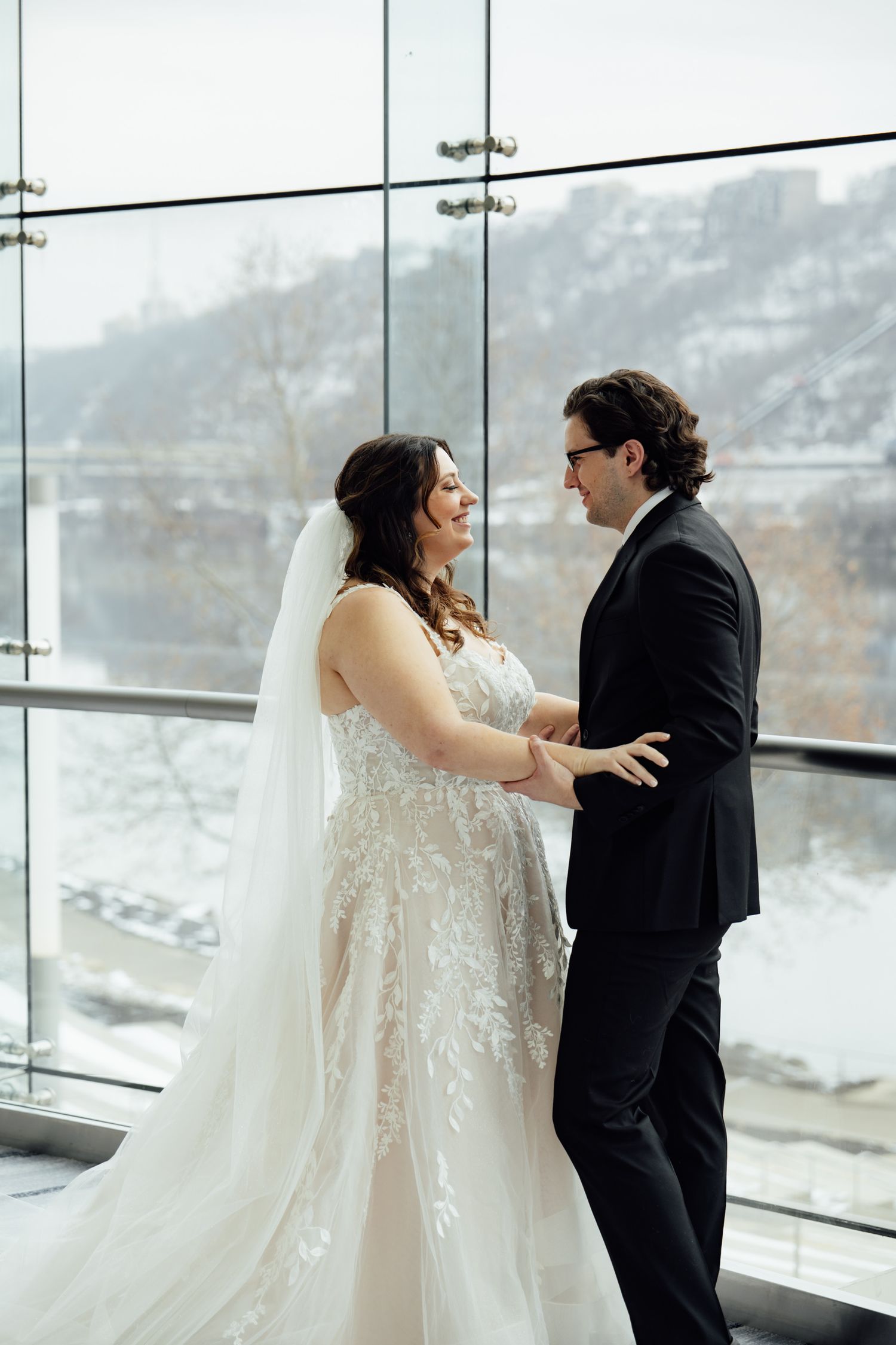 Bride and groom share a romantic first look by floor-to-ceiling windows with a snowy winter landscape backdrop.