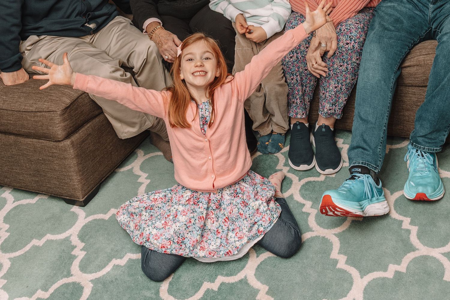 Red-haired girl in pink jacket and floral skirt kneeling on rug with arms outstretched, smiling joyfully with family seated behind.