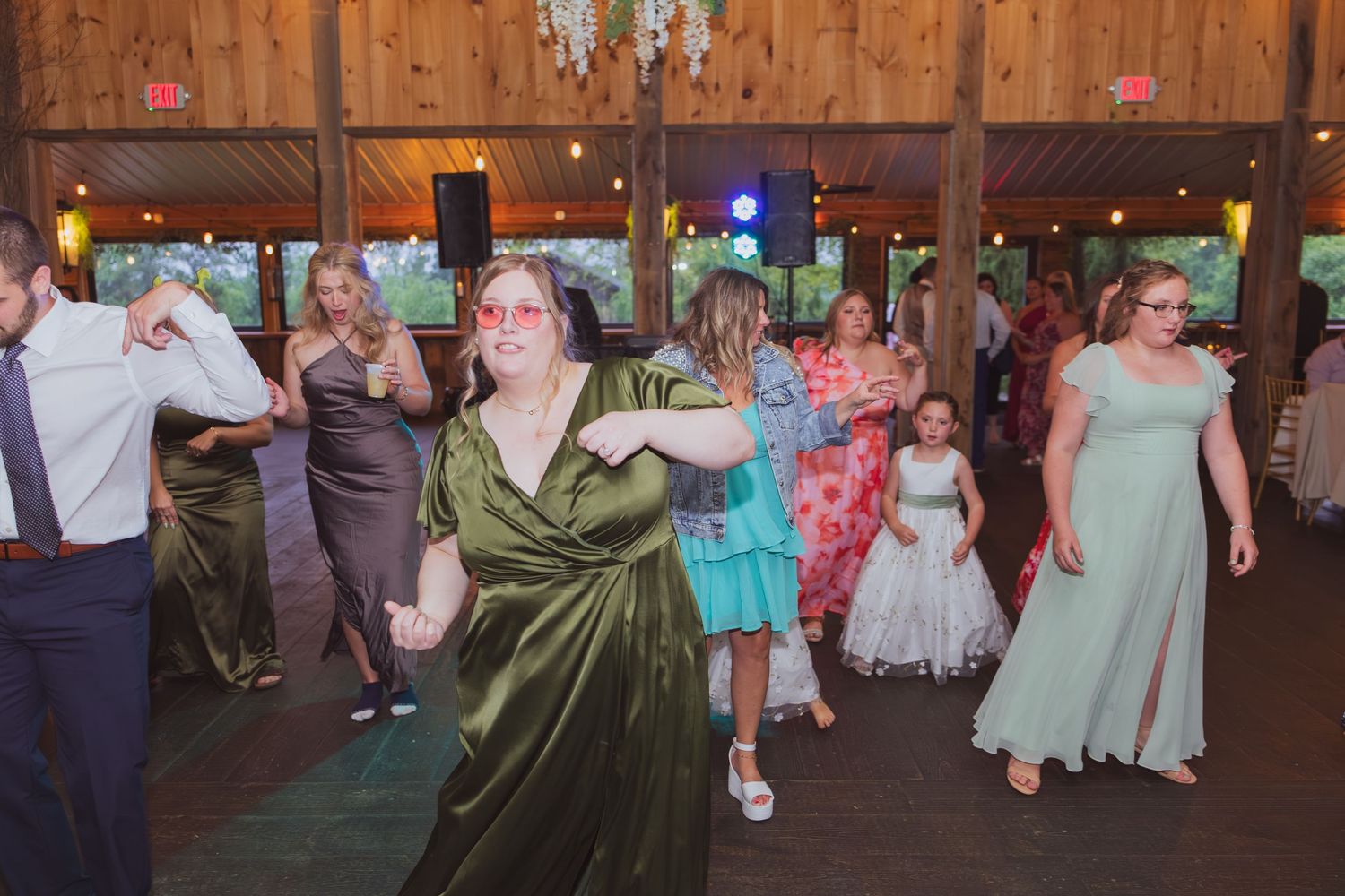 Guests dancing joyfully at a rustic barn wedding reception, wearing formal attire including green and mint gowns.