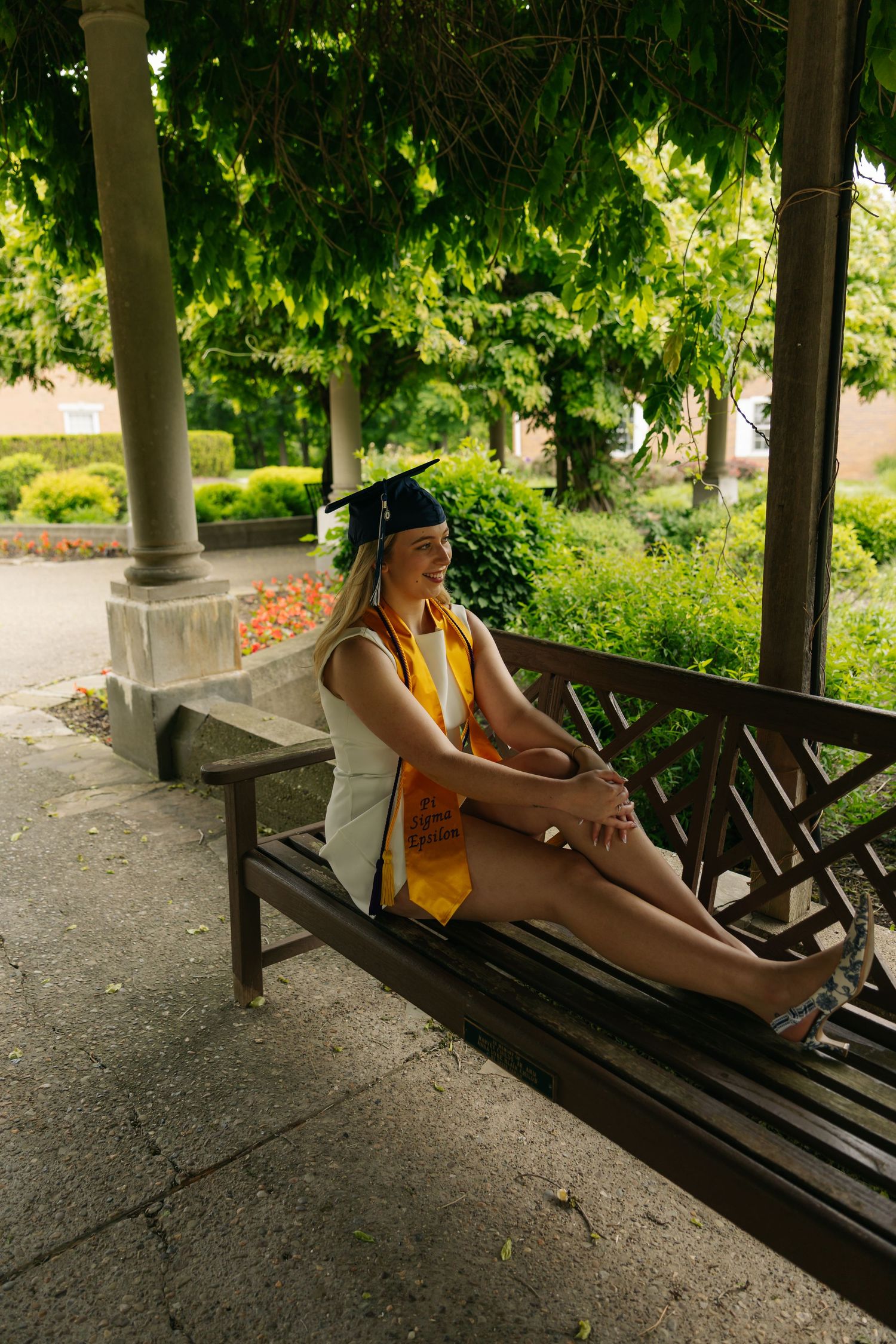 A graduate in cap and gown relaxes on a wooden bench under a green pergola, smiling contentedly.