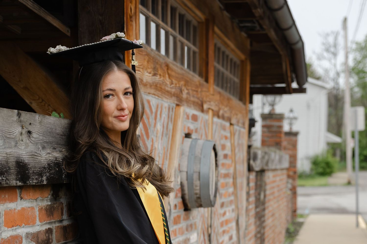 Young woman in graduation cap and gown smiling beside a rustic brick building outdoors.