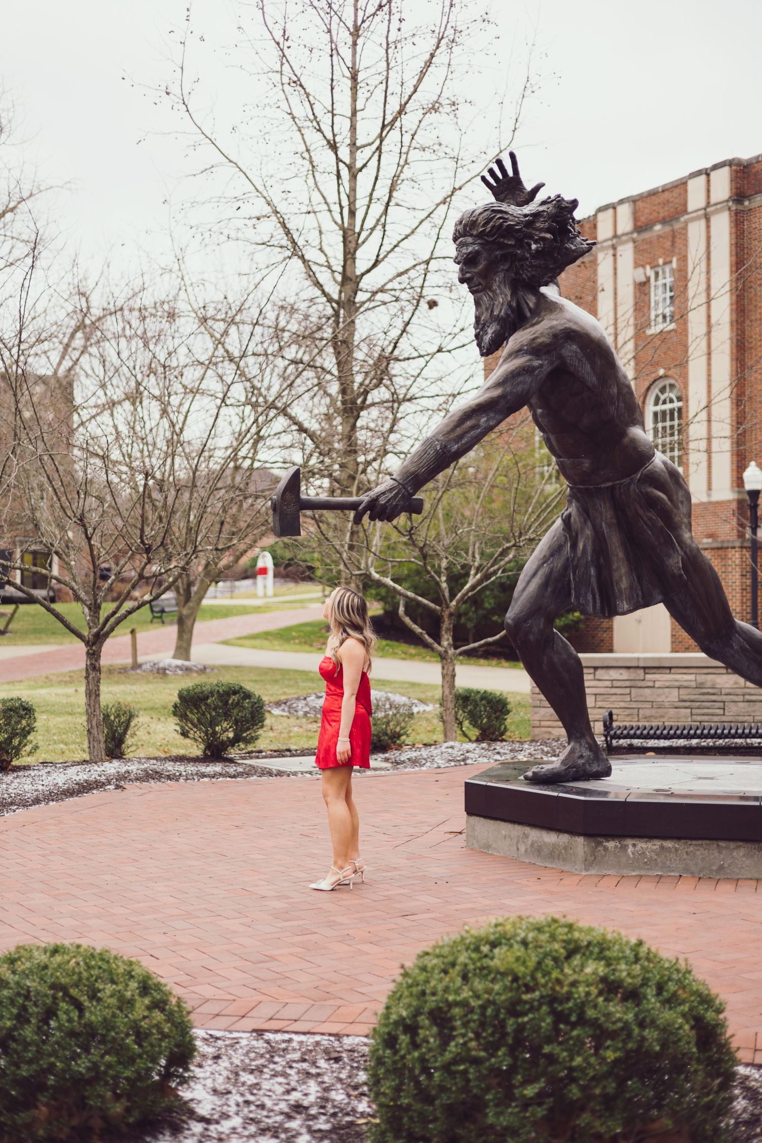 Graduate in red dress poses playfully with campus statue in brick courtyard.