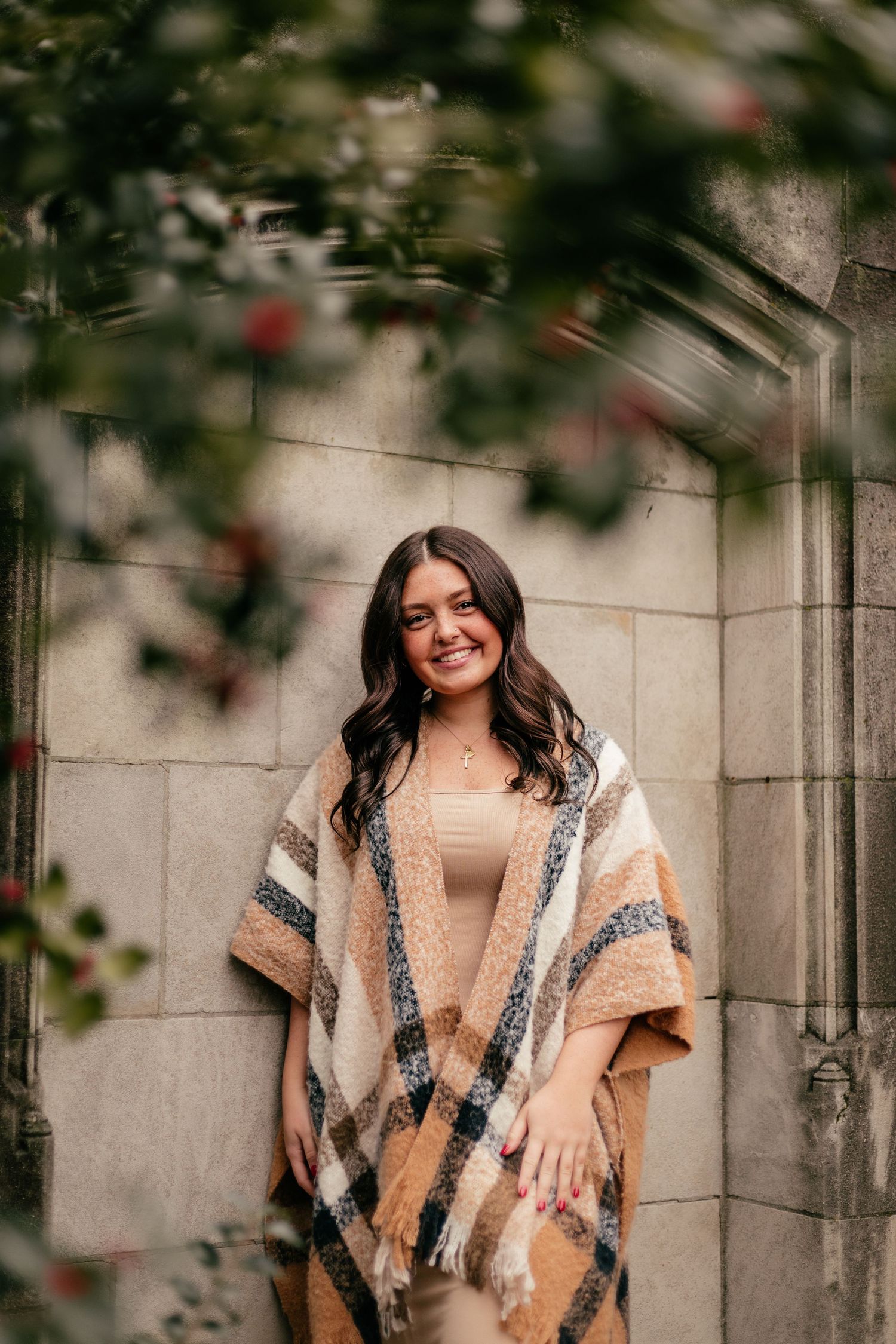 A person wearing a beige and white patterned poncho poses against a stone wall with blurred foliage.