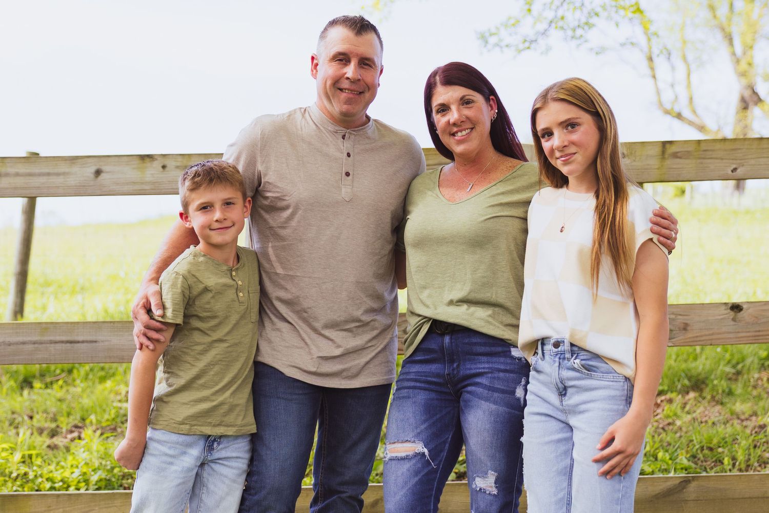 A family poses together outdoors in casual attire standing in front of a wooden fence in a rural setting.