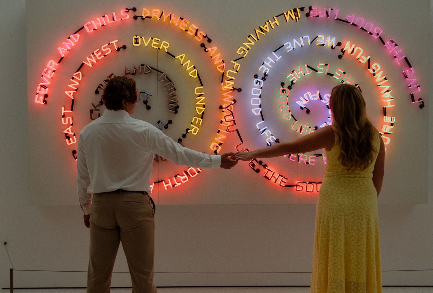 Couple holding hands before a colorful neon light art installation with spiral text in a museum gallery.