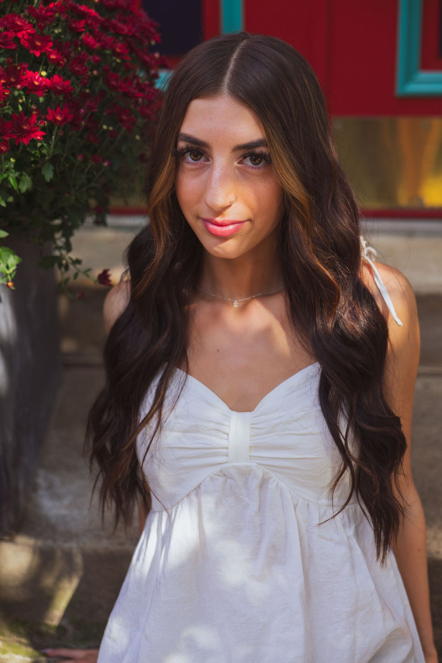Young woman with long dark wavy hair wearing a white sundress, posing outdoors near red flowers.