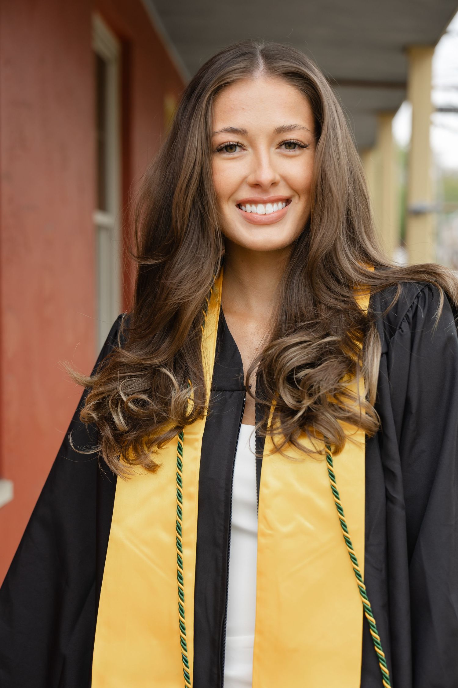 Smiling young woman in black graduation gown with gold stole and green cord, posing outdoors on graduation day.