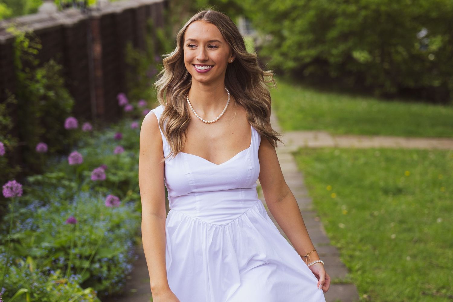 A person wearing a flowing white sundress stands smiling in a garden with purple flowers and greenery in the background.