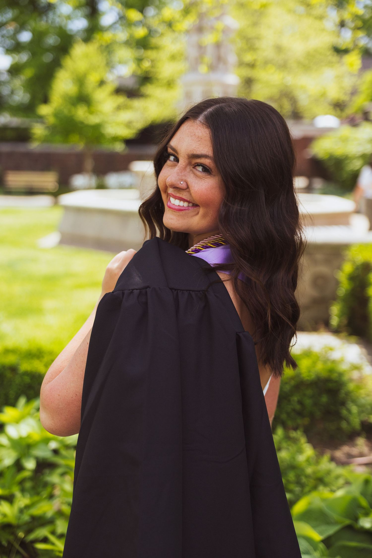 Graduate in black academic regalia poses with a bright smile on sunny spring day with green foliage in background.