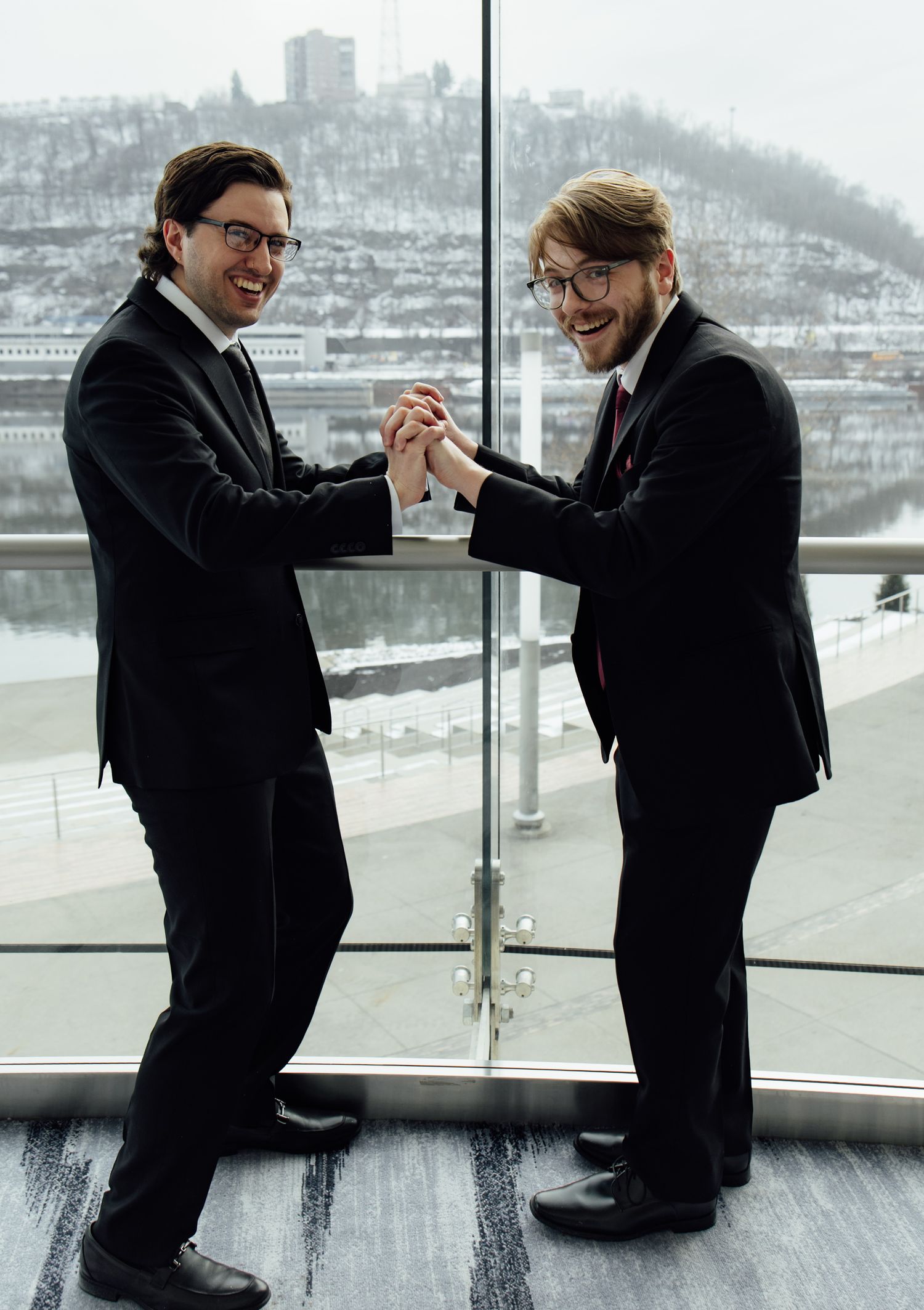Two men in black suits smiling and clasping hands near a glass window with a snowy cityscape backdrop.