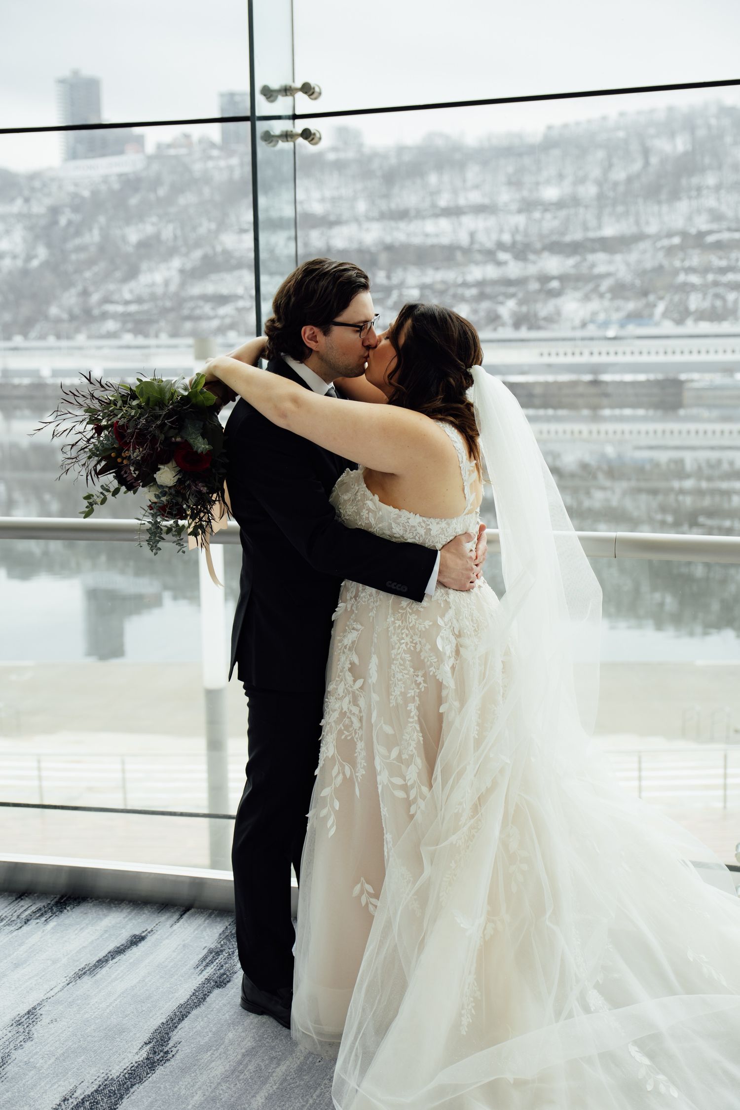 Couple shares first kiss in elegant lace bridal gown and black suit before snowy riverside venue window.