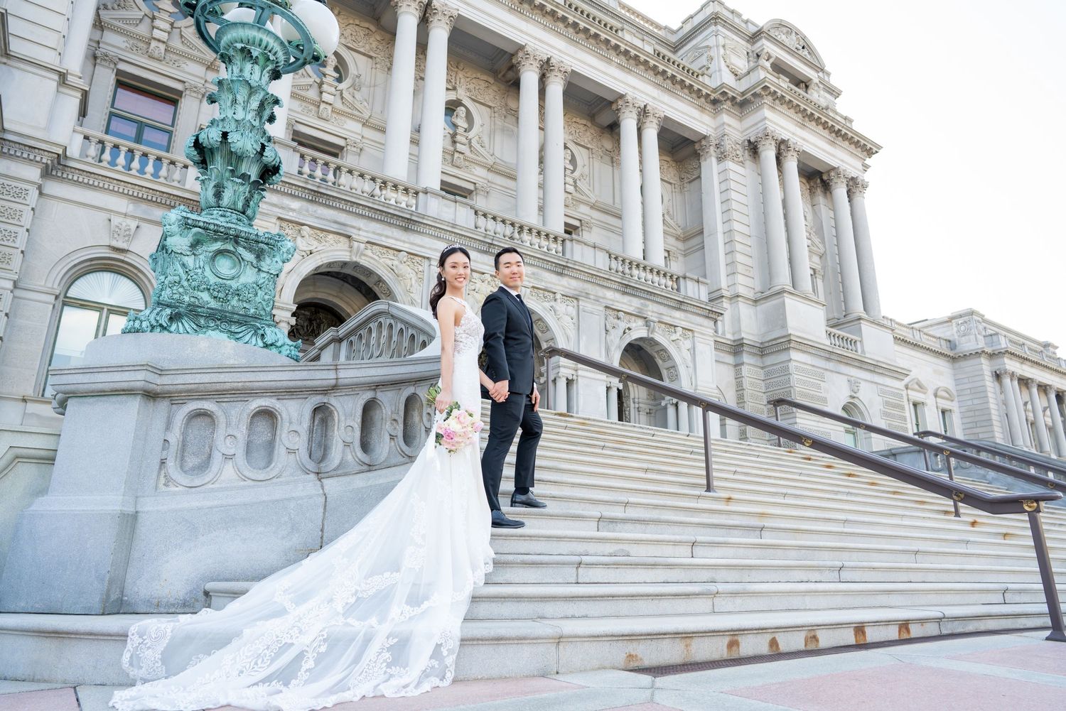 A wedding couple poses on grand stone steps of a historic building with ornate architecture.