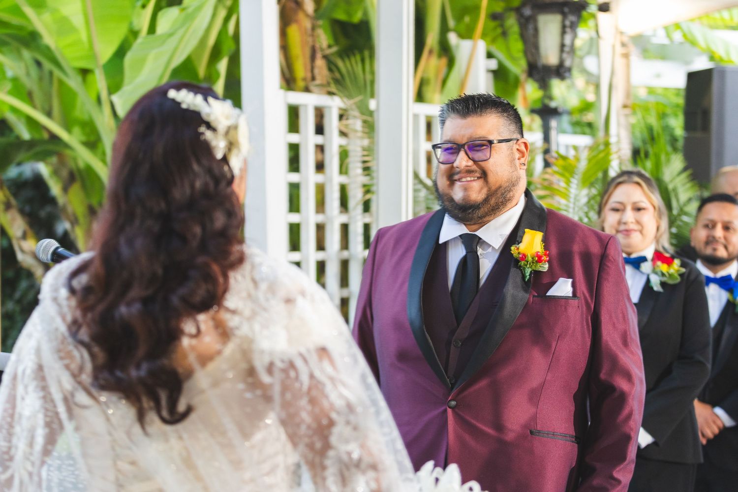 A wedding ceremony takes place outdoors under a white gazebo with tropical greenery.
