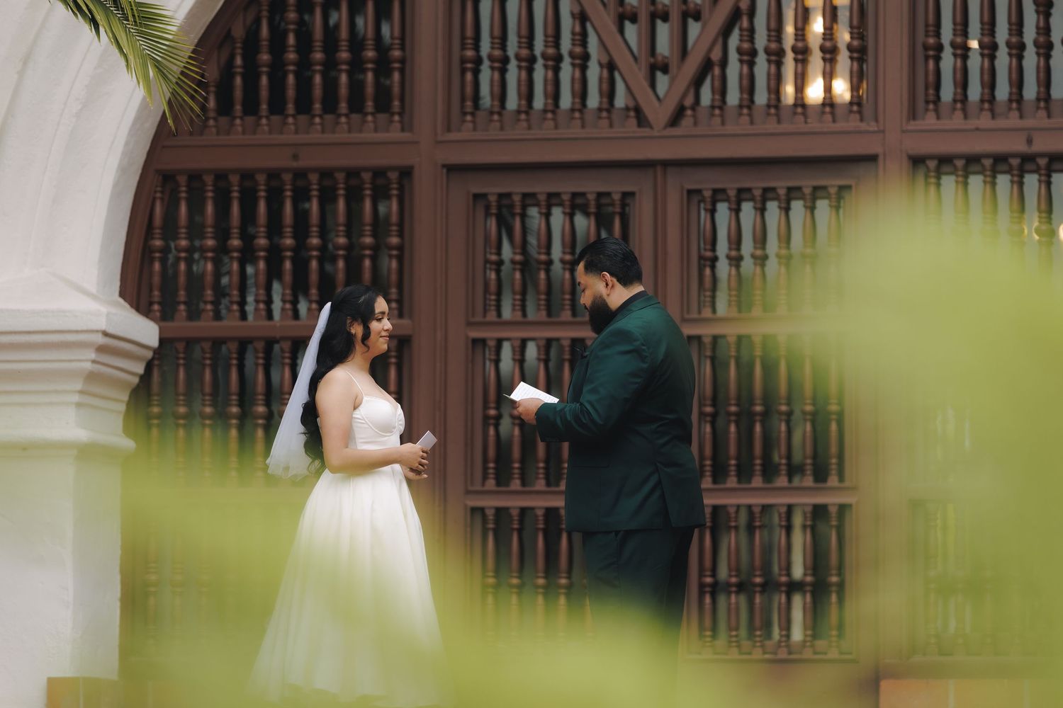 Bride and groom stand in front of ornate wooden bookshelves in a library during their wedding photoshoot.