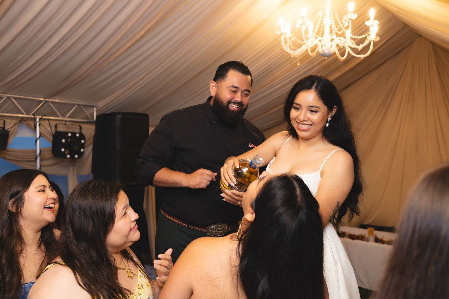 People dancing and celebrating at an indoor wedding reception under a white tent with chandeliers.