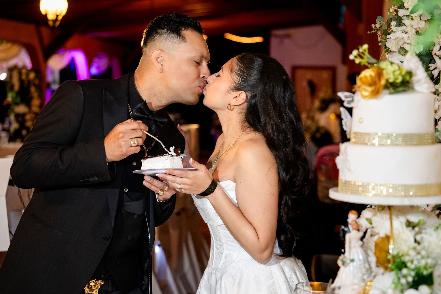 Newlyweds share their first bite of wedding cake at an elegant reception with colorful lighting.