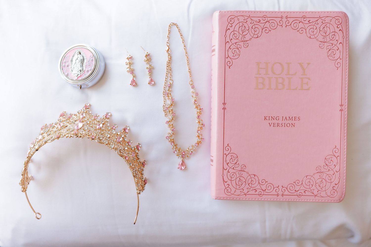 A collection of pink and gold religious items including a Bible, rosary beads, and delicate jewelry arranged on white fabric.
