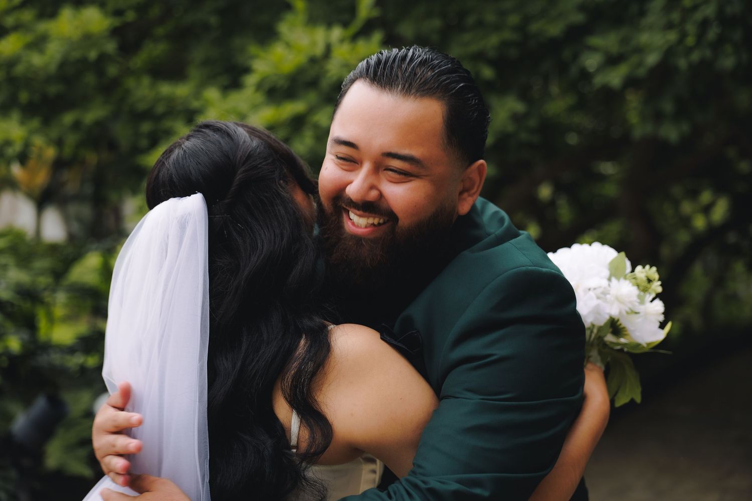 A couple in formal wedding attire embraces and laughs together in a lush outdoor garden setting.